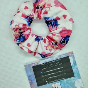 May include: A white and pink scrunchie with a blue cartoon character and pink hearts. The scrunchie is on a white surface with a business card that says "LL Homemade Scrunchies".