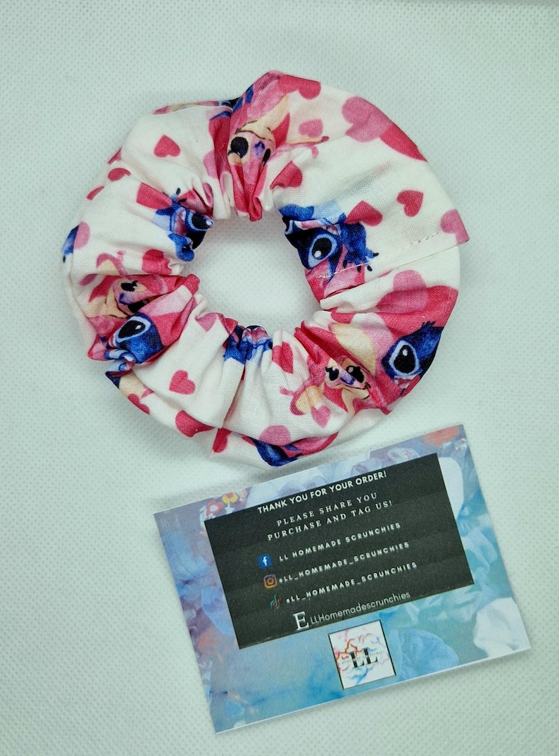 May include: A white and pink scrunchie with a blue cartoon character and pink hearts. The scrunchie is on a white surface with a business card that says "LL Homemade Scrunchies".