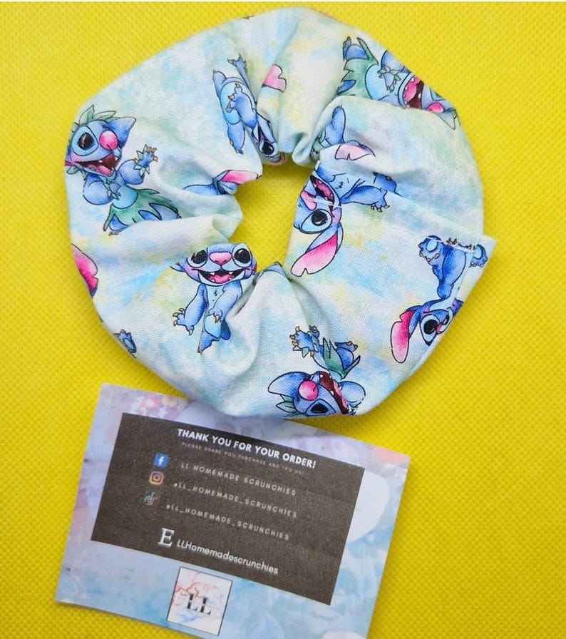May include: A blue and white scrunchie with a cartoon character print. The scrunchie is made of fabric and has a gathered design. The character is blue with pink ears and is wearing a lei. The scrunchie is on a yellow surface. The text on the card reads: "Thank you for your order! #LLHomemadeScrunchies #LLHomemadescrunchies"