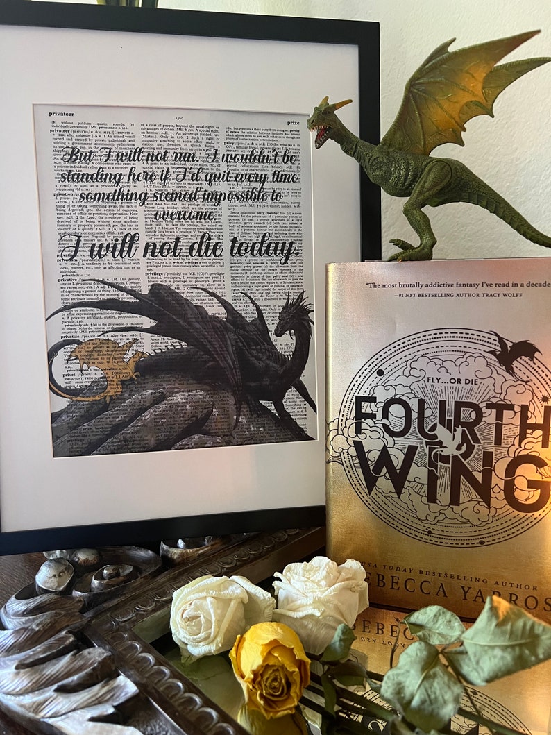 K&ouml;nnte beinhalten: Ein gerahmter Druck mit einem Schwarzwei&szlig;bild eines Drachen mit dem Text "But I will not run. I wouldn't be standing here if I'd quit every time something seemed impossible to overcome. I will not die today."  Ein Buch mit dem Titel "Fourth Wing" von Rebecca Yarros befindet sich im Vordergrund. Der Buchumschlag zeigt einen goldenen Hintergrund mit einer Schwarzwei&szlig;illustration eines Drachen.