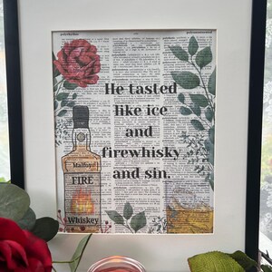 Könnte beinhalten: Gerahmter Kunstdruck mit einem Hintergrund aus einer Wörterbuchseite. Er zeigt eine Whiskeyflaschen-Illustration und den Text "He tasted like ice and firewhisky and sin." Eine rote Rose und grüne Blätter schmücken den Druck.