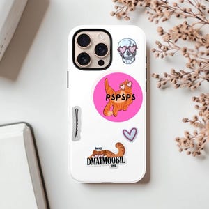 May include: White phone case with multiple stickers. Stickers include a skull wearing heart-shaped sunglasses, a cat with heart-shaped sunglasses inside a pink circle with the text "PSPSPS", a hair pin, a heart, and a cat with the text "in my DMATMOOBIL era".