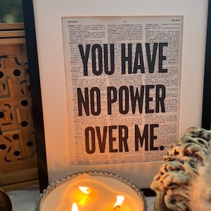 May include: A framed print with the text "YOU HAVE NO POWER OVER ME." printed in black on a vintage dictionary page. The print is displayed on a table with a candle and a decorative sculpture.