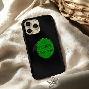 Puede incluir: Funda de teléfono negra con un pop socket verde que dice "Dramione book club magic". La funda del teléfono está sobre una superficie beige con una grulla de origami decorativa.