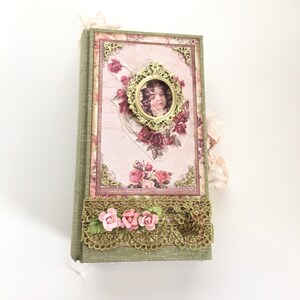 以下が含まれることがあります： ピンクとゴールドのフローラルデザインが施された緑色の布張りのジャーナル。表紙には、花冠をかぶった若い女の子の額縁入り写真が飾られています。ジャーナルにはレースのトリムとピンクの布の花が付いています。