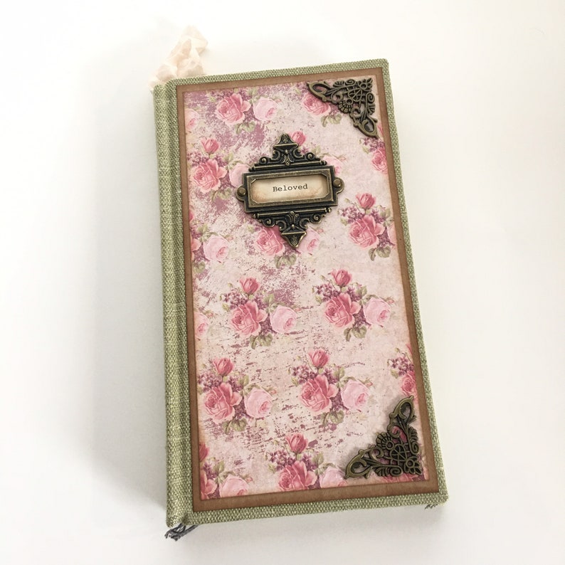 以下が含まれることがあります： ピンクの花柄が施された緑色のリネンカバーのジャーナル。金属製の装飾には「Beloved」と書かれています。
