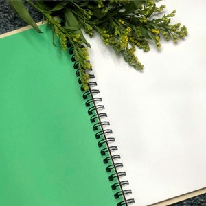 May include: A green and white spiral bound notebook with a black binding. The notebook is open to a blank white page. A sprig of yellow flowers is resting on the notebook.