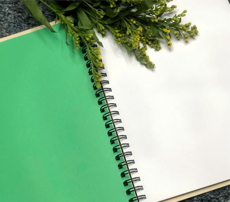 May include: A green and white spiral bound notebook with a black binding. The notebook is open to a blank white page. A sprig of yellow flowers is resting on the notebook.