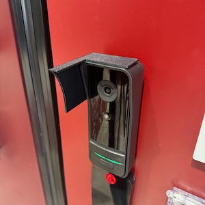 Könnte beinhalten: Eine schwarze Video-Türklingel mit einem Kameraobjektiv, einem grünen Licht und einem roten Knopf. Sie ist auf einer roten Oberfläche montiert und hat eine kleine schwarze Blende über der Kamera.