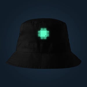 Könnte beinhalten: Ein schwarzer Fischerhut mit einer mittig platzierten, leuchtend hellgrünen Grafik eines Computerprozessors. Das Design zeigt eine quadratische Zentraleinheit mit mehreren Pins, die von allen vier Seiten auf dem schwarzen Stoff abstehen.