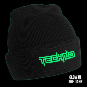 Op de afbeelding: Zwarte beanie met een groene glow-in-the-dark patch met de tekst "TECHNO".