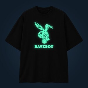Może przedstawiać: Czarny t-shirt z krótkim rękawem z jasną, świecącą na zielono grafiką przedstawiającą sylwetkę głowy królika z wzorami płytek drukowanych oraz okrągłą ikoną. Napis RAVEBOY jest nadrukowany w kolorze jasnej zieleni pod grafiką królika.