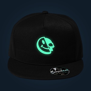 Può includere: Un cappellino snapback nero con una grafica verde chiaro luminosa sul pannello frontale. Il design raffigura un cerchio diviso, con una metà che mostra una faccia sorridente e l'altra metà un teschio. Un adesivo circolare sulla visiera riporta la scritta Snapback.