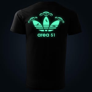 Peut inclure: T-shirt noir avec un motif graphique vert fluorescent. Le motif présente un logo stylisé avec trois ovnis et le texte "area 51". Le graphisme brille dans le noir, créant un effet visuel saisissant.