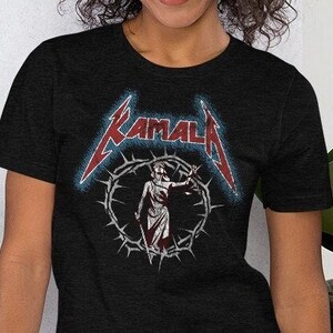 May include: A black t-shirt with a graphic of a woman holding a scale of justice in a crown of thorns. The text "KAMALA" is above the graphic in a stylized font.