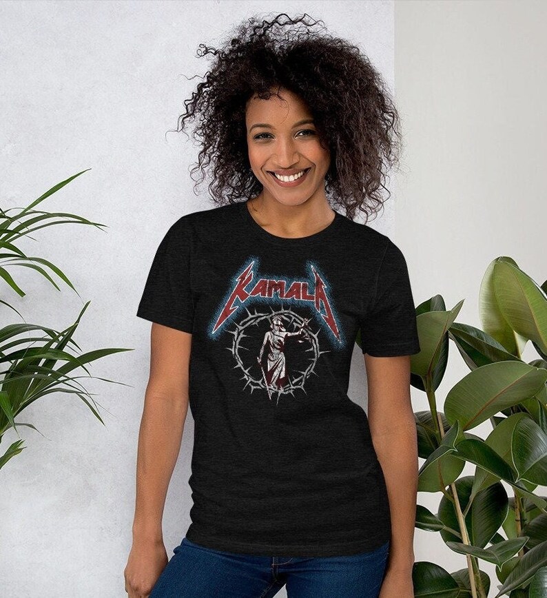 May include: A black t-shirt with a graphic of a woman holding a scale of justice in a crown of thorns. The text "KAMALA" is above the graphic in a stylized font.