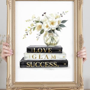 Puede incluir: Impresión enmarcada con un bodegón floral. Un jarrón de cristal con flores blancas se asienta sobre una pila de libros con letras doradas: "LOVE", "GLAM" y "SUCCESS". El marco dorado tiene un diseño ornamentado.