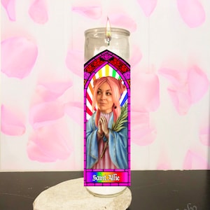 May include: A lit glass candle with a colorful stained-glass design featuring a woman with pink hair and the text "Saint Allie." The candle sits on a white stone, with pink petals in the background.