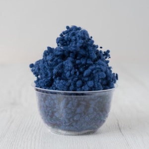 May include: A clear plastic cup filled with a mound of dark blue shredded paper. The paper has a textured, fluffy appearance. The cup sits on a light-colored wooden surface, with a plain white background.