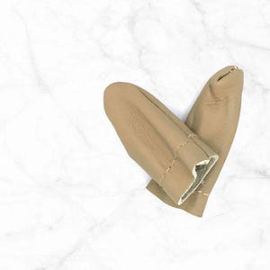 Puede incluir: Dos protectores de dedos de cuero color beige, diseñados para proteger los dedos. Los protectores de dedos están hechos de cuero suave y flexible y están cosidos a los lados. El fondo es un patrón de mármol blanco.
