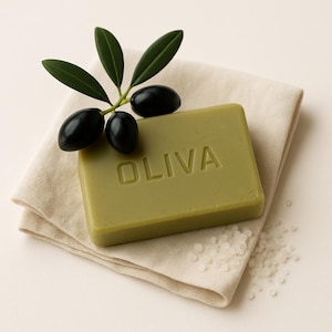 Peut inclure: Un savon vert en barre avec le mot "OLIVA" en relief. Le savon est posé sur un tissu beige avec une branche d'olivier et trois olives noires.