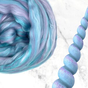 K&ouml;nnte beinhalten: Eine Wirbel aus blauer, t&uuml;rkisfarbener und lavendelfarbener Wolle neben einem hellblauen Einhornhorn mit lila Glitzerakzenten. Das Horn hat ein spiralf&ouml;rmiges Design und ist vor einem wei&szlig;en Hintergrund platziert.