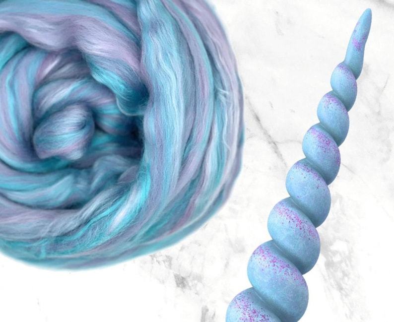 K&ouml;nnte beinhalten: Eine Wirbel aus blauer, t&uuml;rkisfarbener und lavendelfarbener Wolle neben einem hellblauen Einhornhorn mit lila Glitzerakzenten. Das Horn hat ein spiralf&ouml;rmiges Design und ist vor einem wei&szlig;en Hintergrund platziert.
