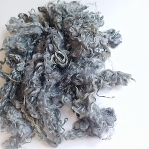 May include: A pile of raw, unspun wool fibres in various shades of grey. The material features a natural, curly, and textured appearance with long, interlocking locks of fibre that vary in depth of colour from light silver to dark charcoal grey.