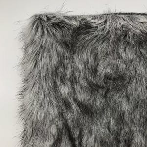 May include: A close-up shot of a rectangular piece of faux fur. The fur is a blend of grey and black, with long, soft strands creating a textured appearance. The edges of the fabric are visible, showing the backing material.