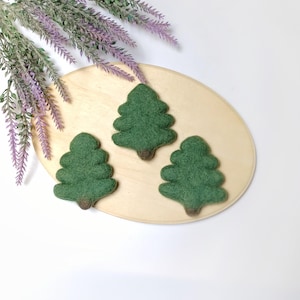 May include: Three green felt Christmas tree ornaments with brown trunks are arranged on a light wooden oval-shaped tray. Lavender sprigs are in the upper left corner. The background is white.