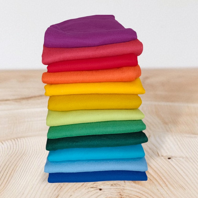 Op de afbeelding: Een verticale stapel van twaalf gevouwen stoffen vierkanten in een kleurenspectrum, waaronder paars, rood, oranje, geel, limoengroen, bosgroen, turkoois, lichtblauw en donkerblauw, netjes gerangschikt in een enkele kolom op een licht oppervlak.