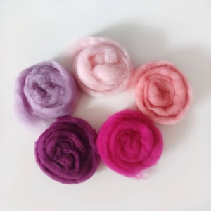 May include: Five balls of wool yarn in shades of pink, purple, and red. The yarn is arranged in a circle on a white surface.