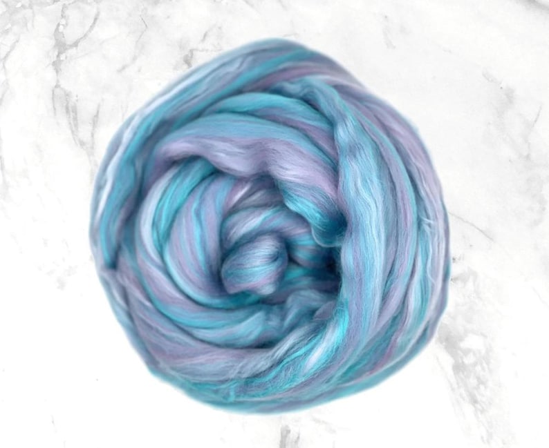 K&ouml;nnte beinhalten: Ein Kn&auml;uel aus Fasern in Blau-, T&uuml;rkis- und Lavendelt&ouml;nen. Die Faser ist eng gewickelt und erzeugt ein wirbelndes Muster. Die Textur wirkt weich und flauschig, geeignet f&uuml;r verschiedene Bastelprojekte.