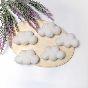 Może przedstawiać: Pięć puszystych, białych filcowych dekoracji w kształcie chmur ułożonych na jasnym drewnianym owalu. Chmury są miękkie i zaokrąglone, idealne do dekoracji pokoju dziecięcego lub projektów rzemieślniczych. Fioletowe gałązki lawendy w tle.