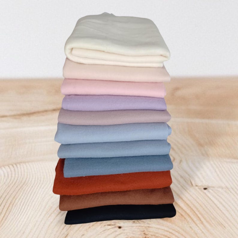 Op de afbeelding: Een verticale stapel gevouwen stukken stof in een kleurverloop, beginnend met wit aan de bovenkant, overgaand in tinten roze, lavendel, blauw en roest, en eindigend met een donkerbruin en zwart stuk onderaan de stapel.
