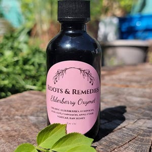 Könnte beinhalten: Eine schwarze Glasflasche mit Tropfer, beschriftet mit "Roots & Remedies Elderberry Oxymel". Das rosa Etikett listet Zutaten wie Holunderbeeren und rohen Honig auf. Ein Zweig grüner Blätter liegt vor der Flasche. Die Flasche ist 59 ml.