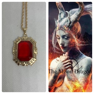Pode incluir: Um colar com pingente dourado com uma grande pedra preciosa vermelha facetada retangular. O pingente é colocado em uma moldura dourada decorativa. O colar é exibido ao lado de uma imagem de um personagem de fantasia com o texto "The Moon Dragon".