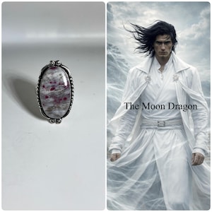 May include: Oval-shaped silver ring with a large, speckled gemstone. The stone features a mix of gray and white with red inclusions. The ring has a decorative border. The image also includes a person in a white coat with the text "The Moon Dragon".