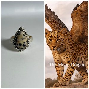 May include: A silver ring with a teardrop-shaped stone featuring a speckled pattern of black and cream. The ring is set against a white background. The image also includes a fantasy illustration of a leopard with wings, and the text "The Moon Dragon".