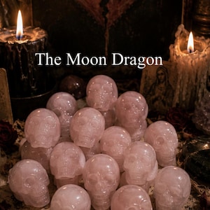 以下が含まれることがあります： ピンク色のクリスタルスカルの置物のコレクション。「The Moon Dragon」の文字がスカルの上に表示されています。背景には、キャンドル、ドライフラワー、壊れたハートの絵画が含まれています。