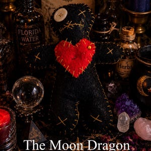 May include: A black fabric doll with a red heart patch, a white button eye, and yellow stitched details. The doll is surrounded by candles, glass bottles labeled Florida Water and Herbs, crystals, and a sign reading Asé By Sister Selena.