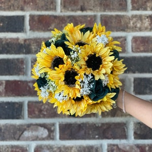 May include: A bouquet of yellow sunflowers with dark green roses and white baby's breath.
