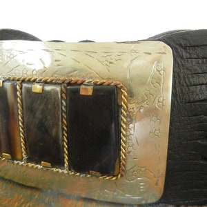 May include: A black leather belt with a silver buckle featuring a decorative design and three rectangular inlays of brown and black horn. The buckle is accented with a gold rope-like border.
