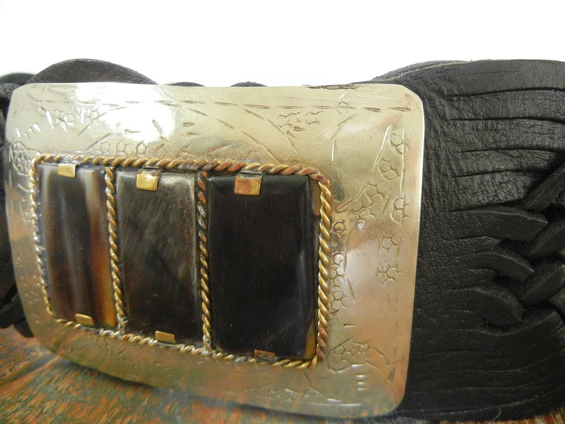 May include: A black leather belt with a silver buckle featuring a decorative design and three rectangular inlays of brown and black horn. The buckle is accented with a gold rope-like border.