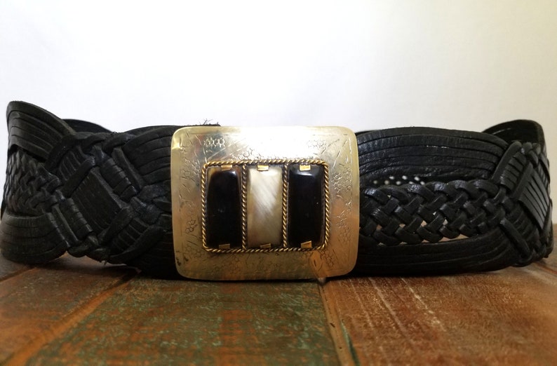 May include: A black leather belt with a braided design and a silver buckle with three inlaid stones. The stones are black, white, and black.