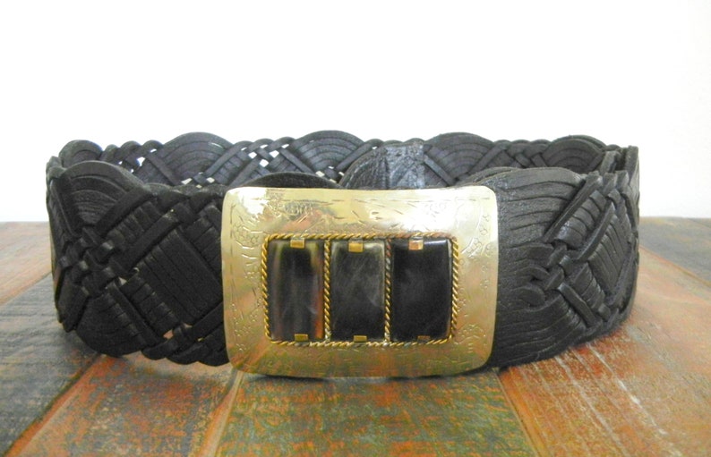 May include: A black leather belt with a woven design and a silver buckle with three black rectangular stones.
