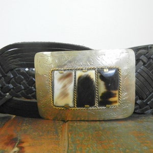 May include: A black leather braided belt with a silver buckle featuring a decorative design and three rectangular horn inlays.