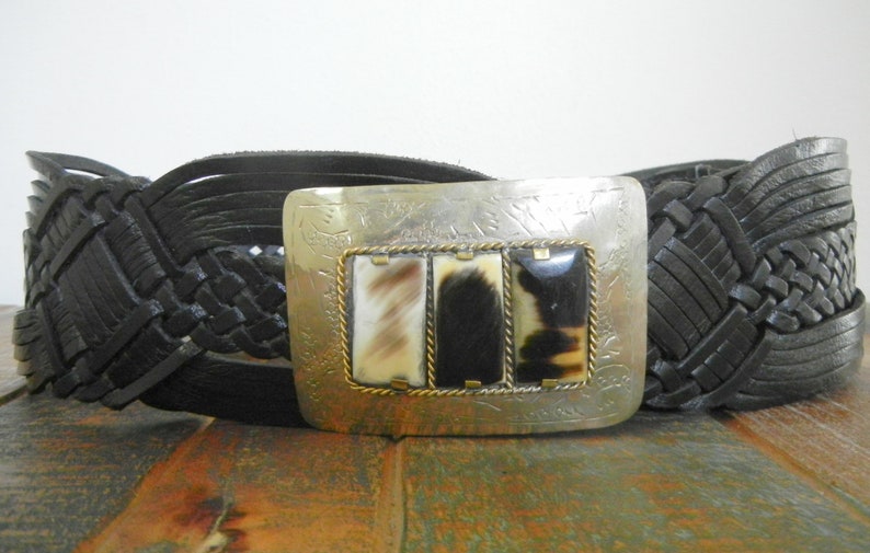 May include: A black leather braided belt with a silver buckle featuring a decorative design and three rectangular horn inlays.