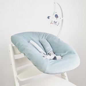 May include: A light blue baby seat cushion with a safety harness, placed on a white wooden high chair. A decorative mobile with a star-shaped design and woodland illustrations hangs above the seat.