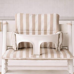 May include: A white wooden high chair with a beige and white striped cushion and backrest. The chair has a white tray and a black footrest. The high chair is designed for infants and toddlers.